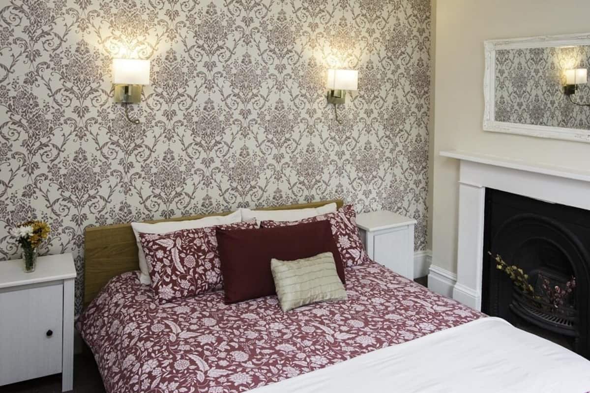 Un dormitorio con papel tapiz estampado, dos lámparas de noche, una cama con colcha floral, una mesita de noche con un florero y una chimenea.
