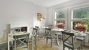 A dining area with a white table and chairs, a small white cabinet, and two windows with plants. - Primrose Guest House (London)
