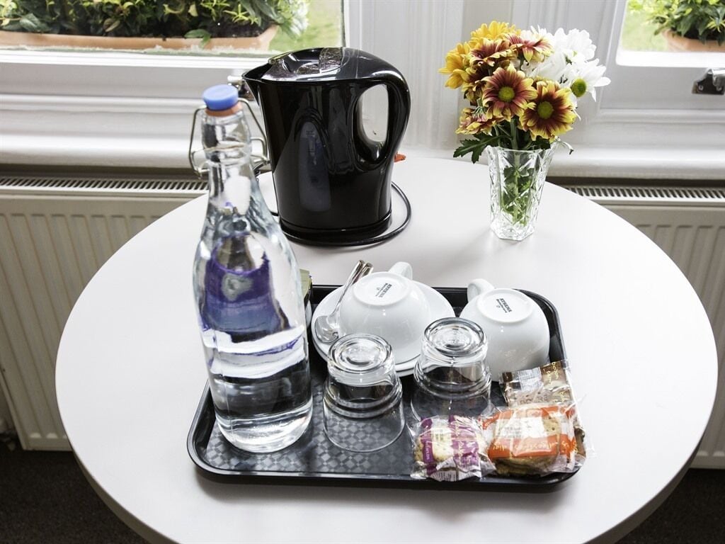 A tray with a water bottle, a black coffee maker, white teapots, glass cups, and a small bag of snacks on a white table near a window with flowers.