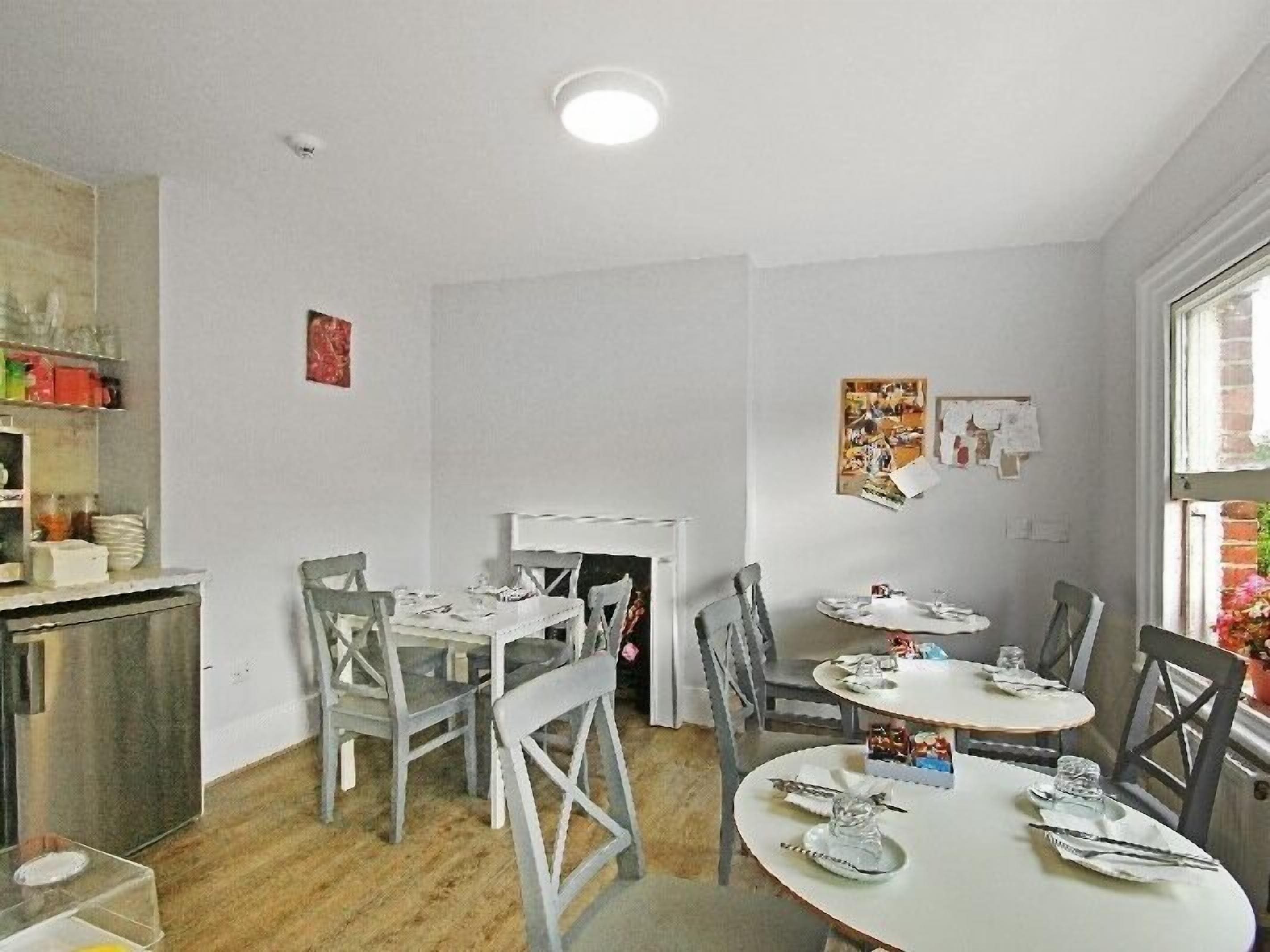 Un comedor con una mesa blanca dispuesta para cuatro personas, rodeada de sillas grises. A la izquierda hay un pequeño armario blanco y a la derecha, una ventana.