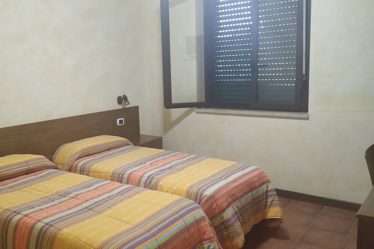 A room with two beds, each with striped bedding, a wooden headboard, a small bedside table, and a window with blinds.