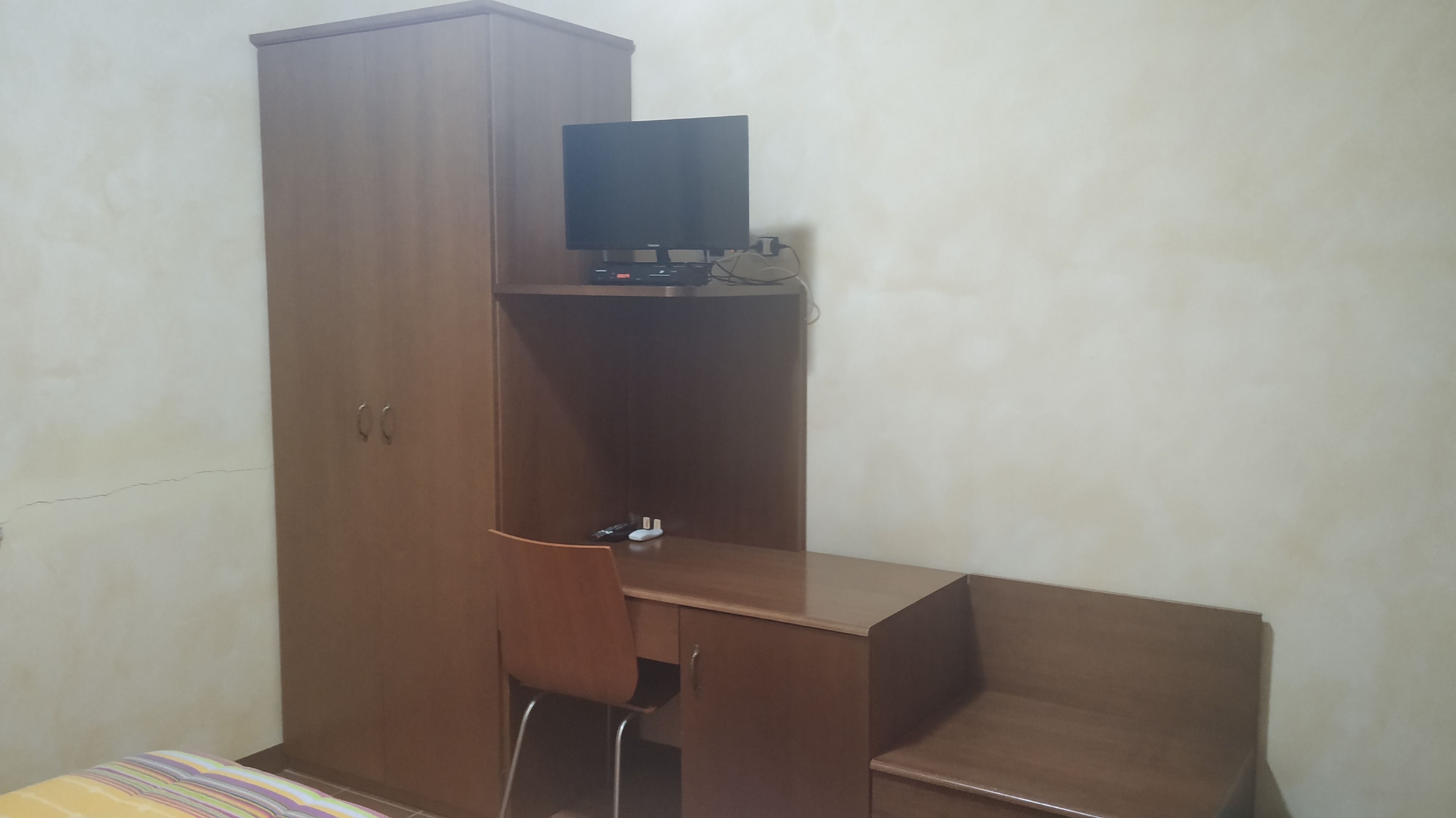 A room with a tall wooden wardrobe, a desk with a computer, and a chair.