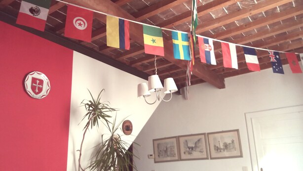 A room with a wooden ceiling, a red wall, and a string of flags. There are framed pictures on the wall and a plant. - La Maison del Corso B&B (Pisa)