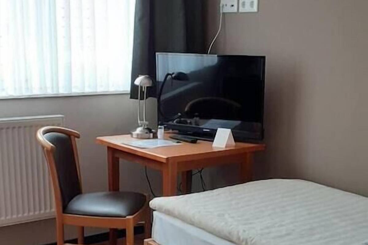 A hotel room with a bed, a desk with a computer and a chair, a window with curtains, and a radiator.