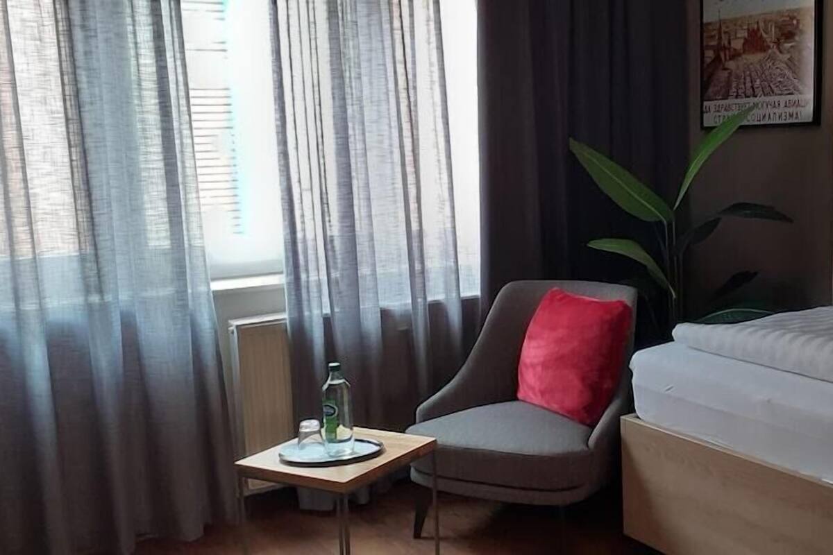A hotel room with a bed, a chair with a red pillow, a small table with a bottle, and a plant.