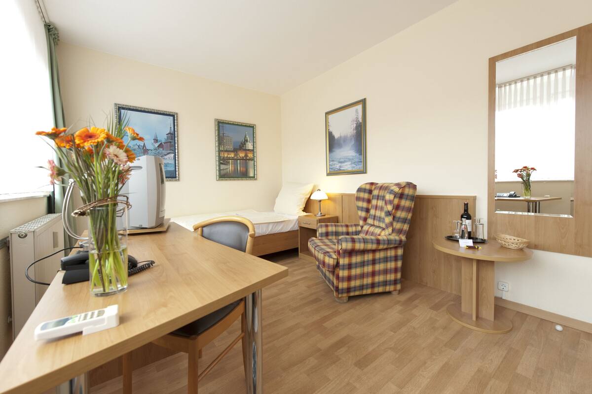 A hotel room with a wooden desk, a plaid armchair, a bed, and two framed pictures on the wall.