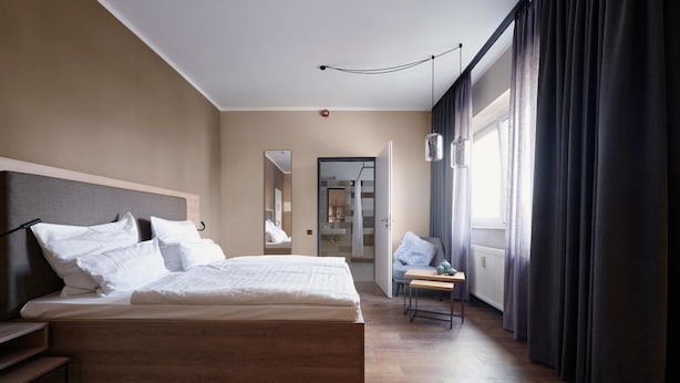 A modern hotel room with a large bed, a small table, and a view of a cityscape through the window. - Hotel Marco Polo (Münster)