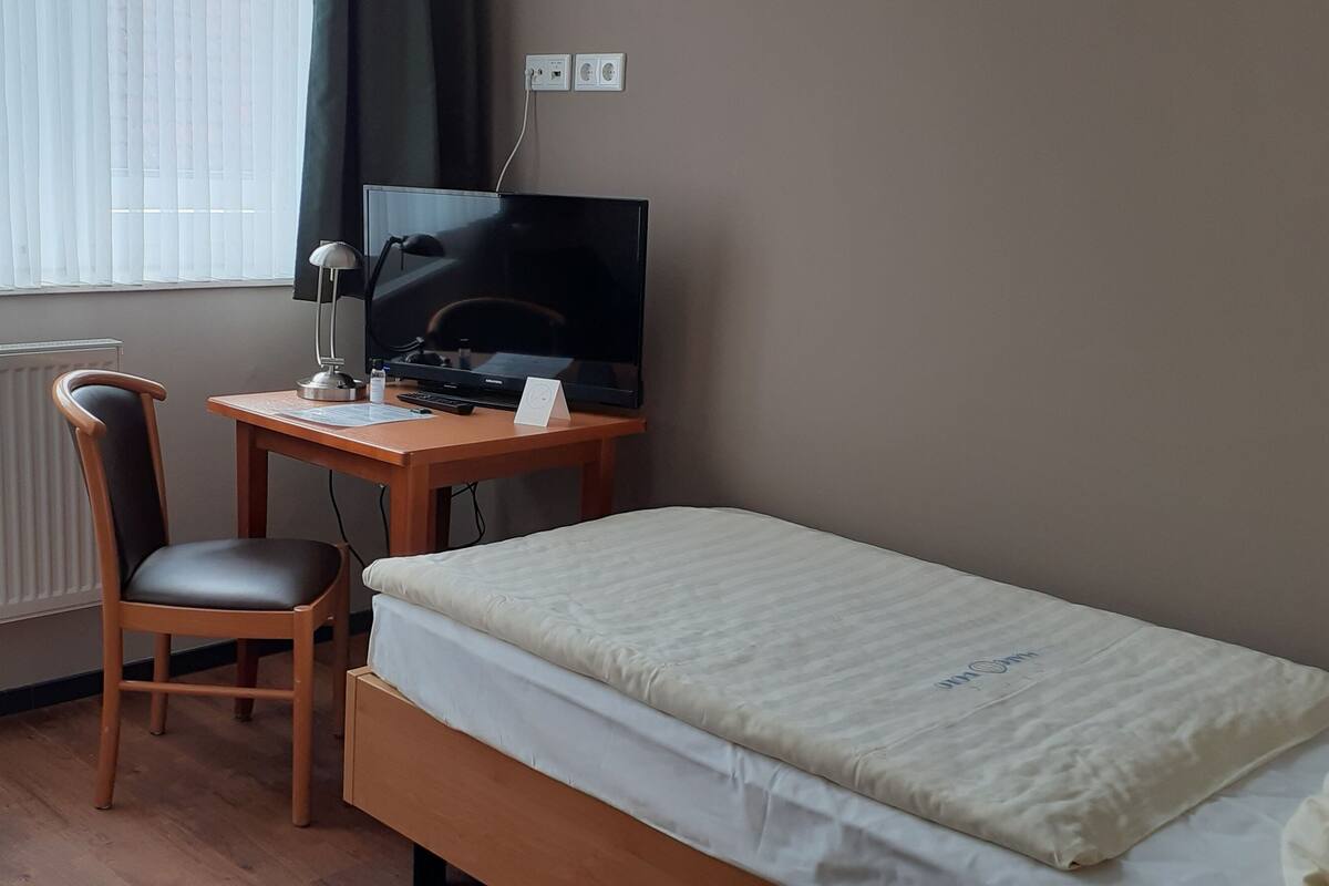 A small hotel room with a single bed, a desk with a computer and a chair, a radiator, and a window with blinds.