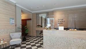 A hotel reception area with a checkered floor, a reception desk, a seating area with a chair, and a potted plant. - Hotel Marco Polo (Münster)