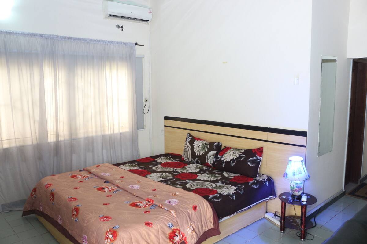 Un dormitorio con cama, lámpara de noche y ventana con cortinas.
