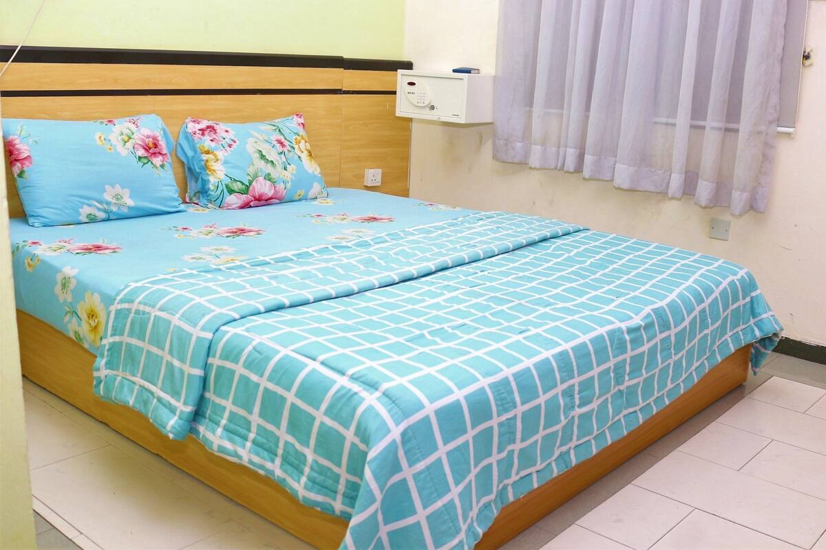 Una cama con ropa de cama floral y a cuadros, un cabecero de madera, una mesita de noche con toma de corriente y una ventana con cortina translúcida.