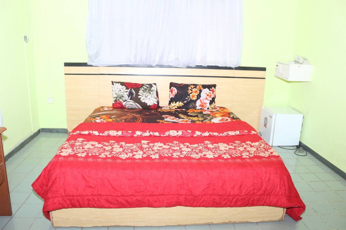 Una cama con ropa de cama floral, una cortina blanca y un pequeño dispositivo blanco en la pared.