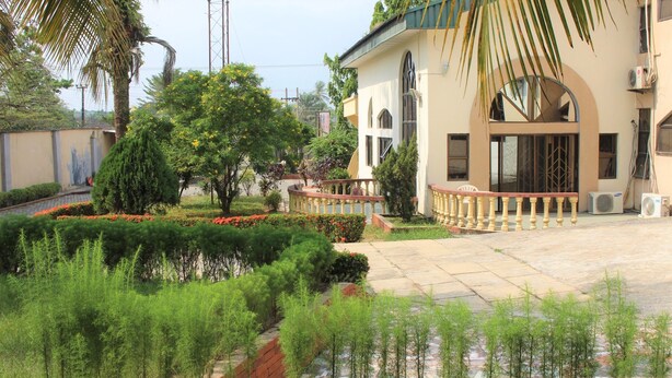 A building with a curved balcony, a palm tree, and a well-maintained garden with red flowers and green shrubs. - Tourist Castle Hotel and Suites (Calabar)
