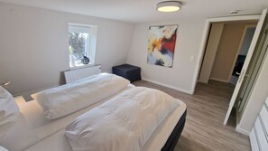 Ein Schlafzimmer mit zwei Betten, einem Fenster, einem Heizkörper, einem Bild und einer Tür zu einem anderen Raum.