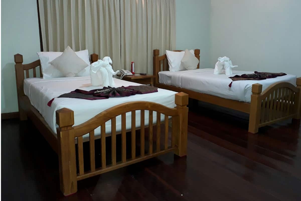 Two wooden beds with white linens and pillows, a wooden nightstand with a book and a red object, and a large window with curtains.
