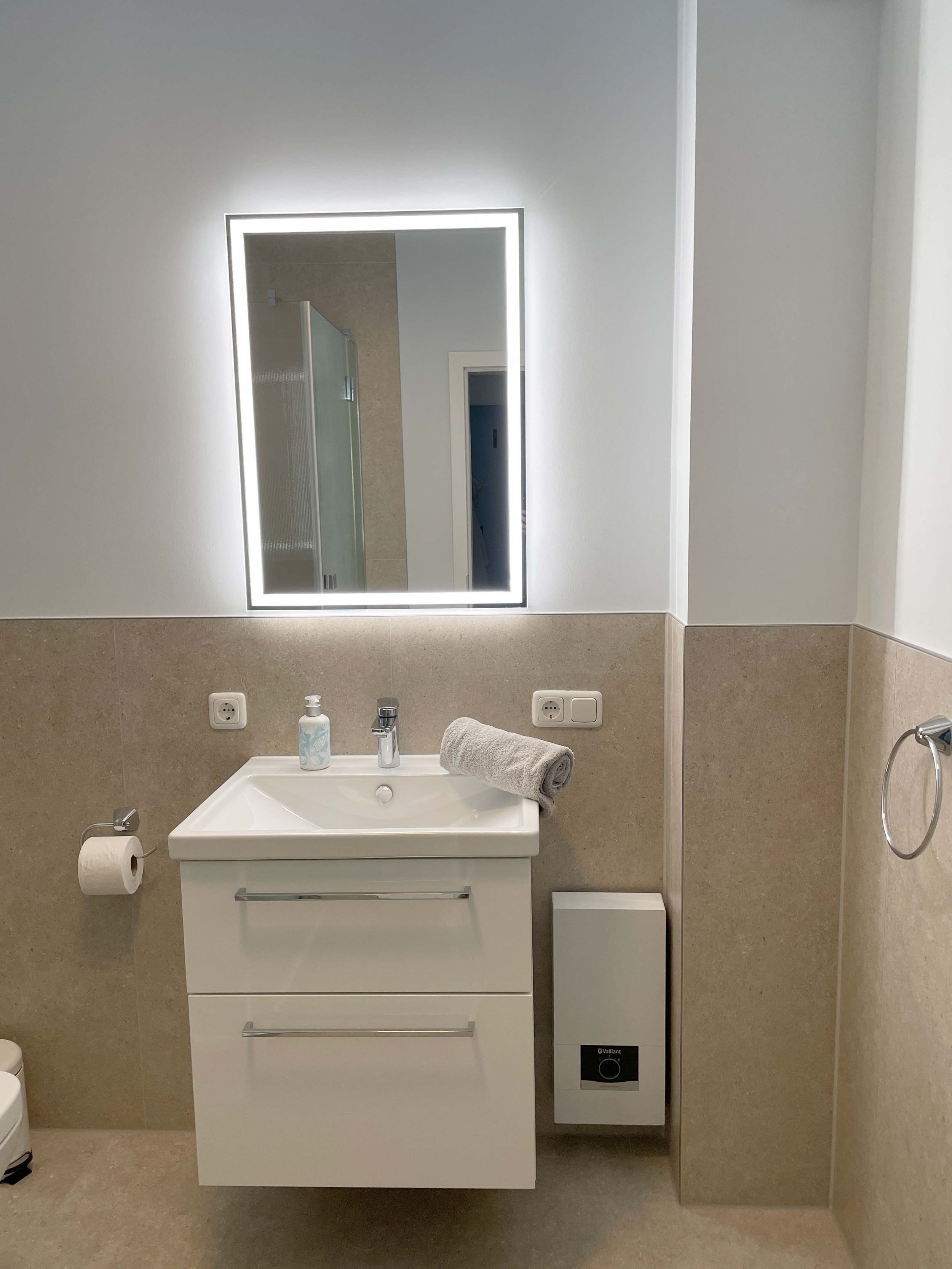 Ein Badezimmer mit weißem Waschbecken, einem Spiegel mit integrierter Beleuchtung, einem Handtuchhalter, einem Toilettenpapierhalter und einem Händetrockner.