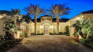 A well-lit, paved entrance to a building with a central archway, flanked by palm trees and a brick pathway. - Escape to Legends - Pool, Games & Amazing Mountain Views in PGA West #067651 5br (La Quinta)