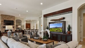 A spacious living room with a large flat-screen TV, comfortable seating, a fireplace, and a kitchen in the background. - Escape to Legends - Pool, Games & Amazing Mountain Views in PGA West #067651 5br (La Quinta)