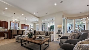 A spacious living room with a large sofa, a coffee table, and a dining area with wooden cabinets and a kitchen in the background. - Escape to Legends - Pool, Games & Amazing Mountain Views in PGA West #067651 5br (La Quinta)
