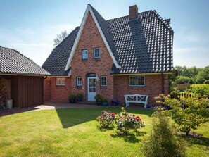 A brick house with a steeply pitched roof, a front door, and a small front yard with a bench and potted plants.
