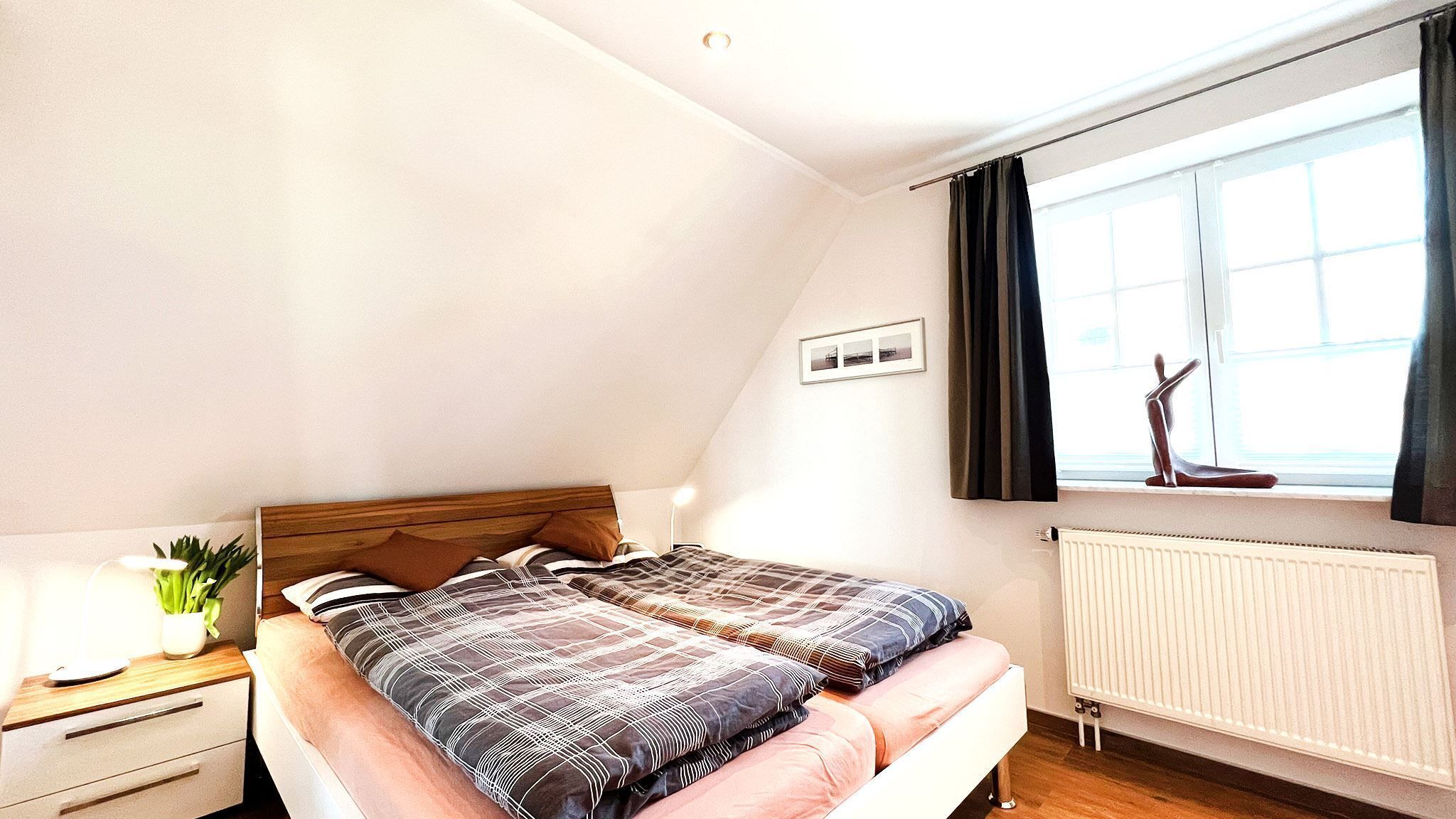 Ein Schlafzimmer mit Bett, Nachttisch, Lampe, Fenster und Heizkörper.