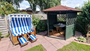 A wooden deck with a blue and white striped beach chair, a small wooden table, and a gazebo with a tiled roof.