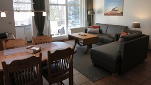 A living room with a sofa, a dining table, chairs, a lamp, and a painting on the wall. - 035-02 (Room category) - House Calidris (Sankt Peter-Ording)