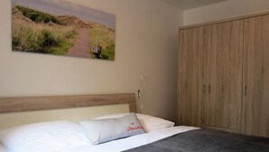 A bedroom with a bed, a wooden wardrobe, a wall-mounted picture of a landscape, and a bedside table with a lamp. - 035-02 (Room category) - House Calidris (Sankt Peter-Ording)
