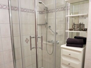 Une douche en verre avec des accessoires chromés, un mur recouvert de carreaux blancs agrémenté d’une bordure décorative, et un meuble-lavabo blanc avec un miroir et une étagère où sont posées des serviettes.