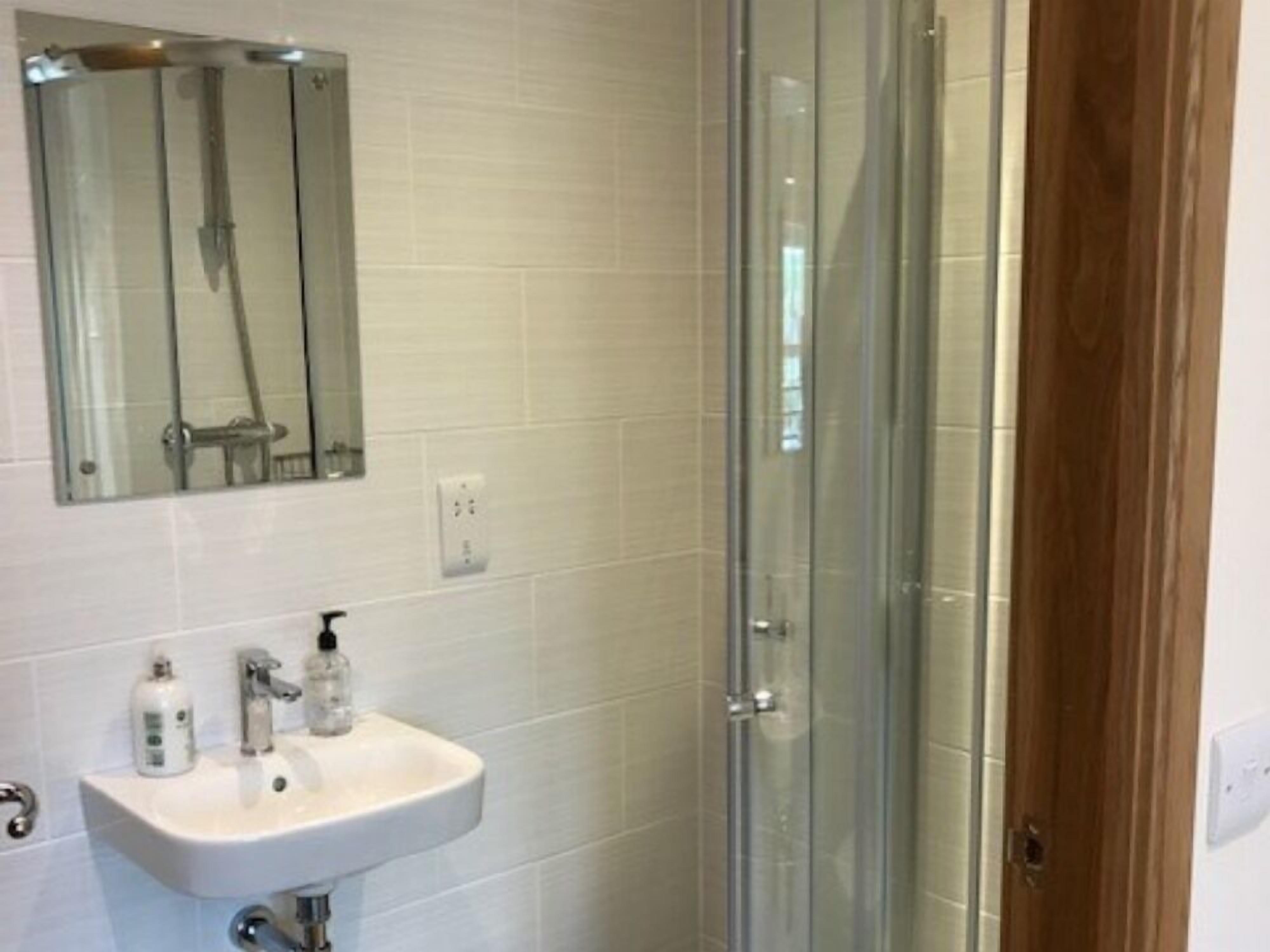 A bathroom with a glass shower enclosure, a white sink with a mirror above it, and a wooden door.