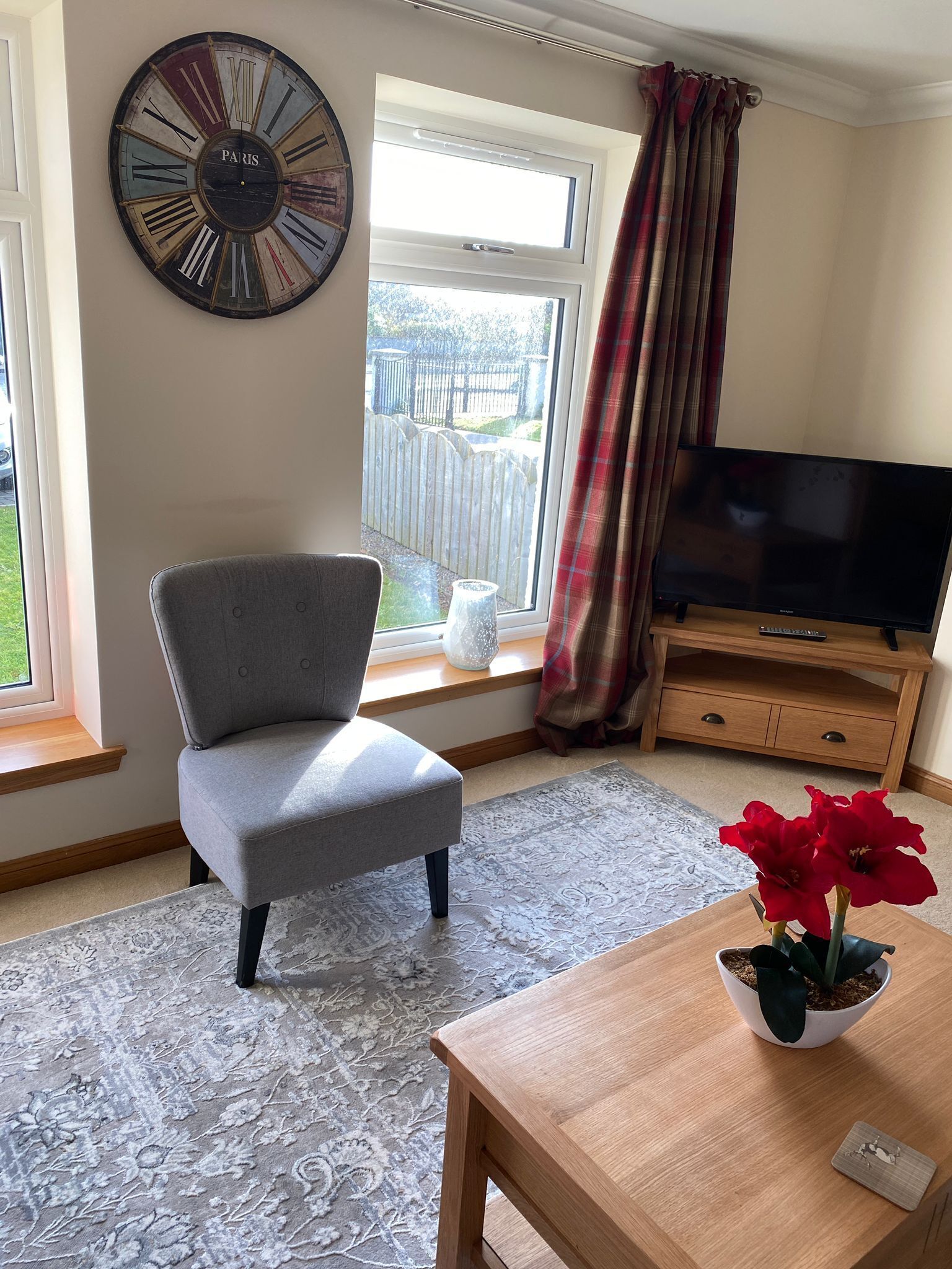 A living room with a grey armchair, a wooden coffee table, a TV on a wooden stand, a large clock on the wall, and a window with a view of a fence.
