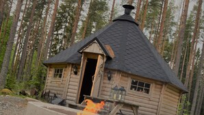 Une cabane en rondins avec une cheminée, un foyer à l’extérieur et une forêt en arrière-plan.