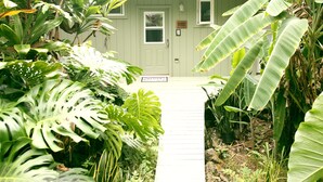 Un sendero de madera que lleva a una cabaña con puerta y ventanas, rodeada de exuberante vegetación tropical.