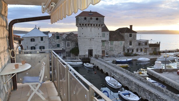 A balcony with a table and chairs overlooking a harbor with boats and historic stone buildings. - Apartments Kastilac (Kastela)