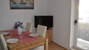 A dining area with a wooden table set for four, a flat-screen TV on a stand, and a painting above the table. - Apartments Kastilac (Kastela)
