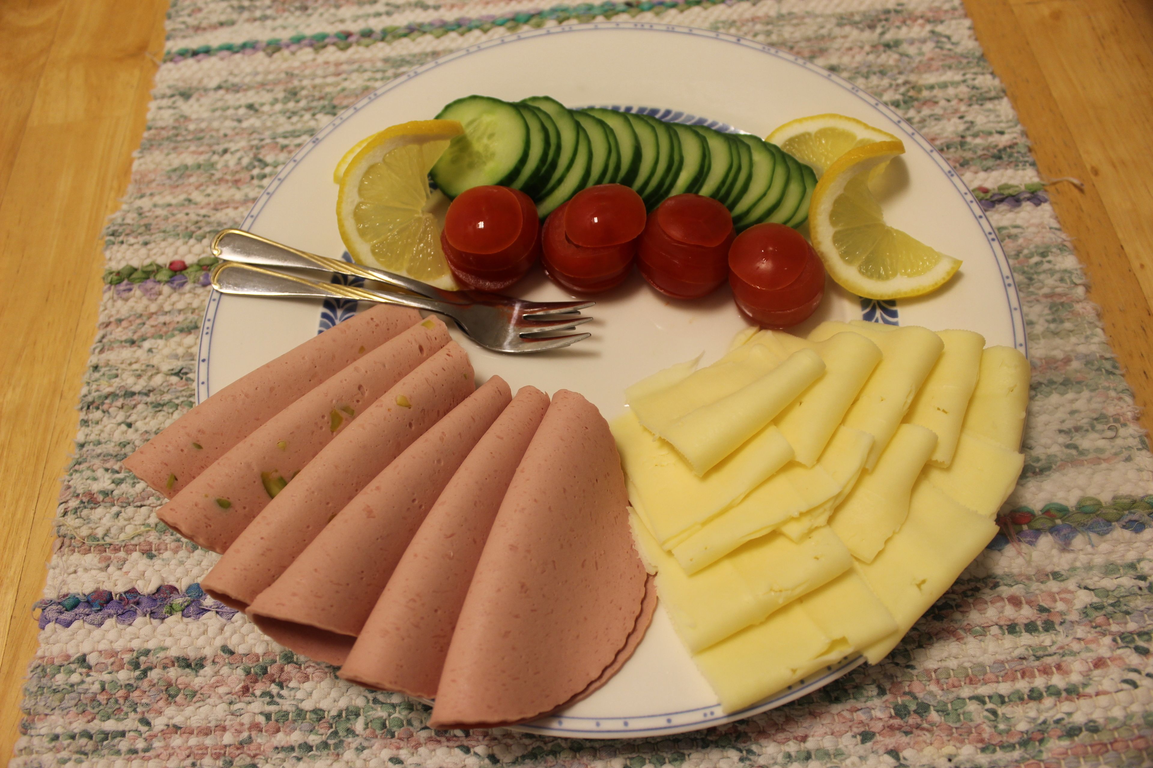 A plate with sliced cucumbers, cherry tomatoes, lemon slices, ham, and cheese.