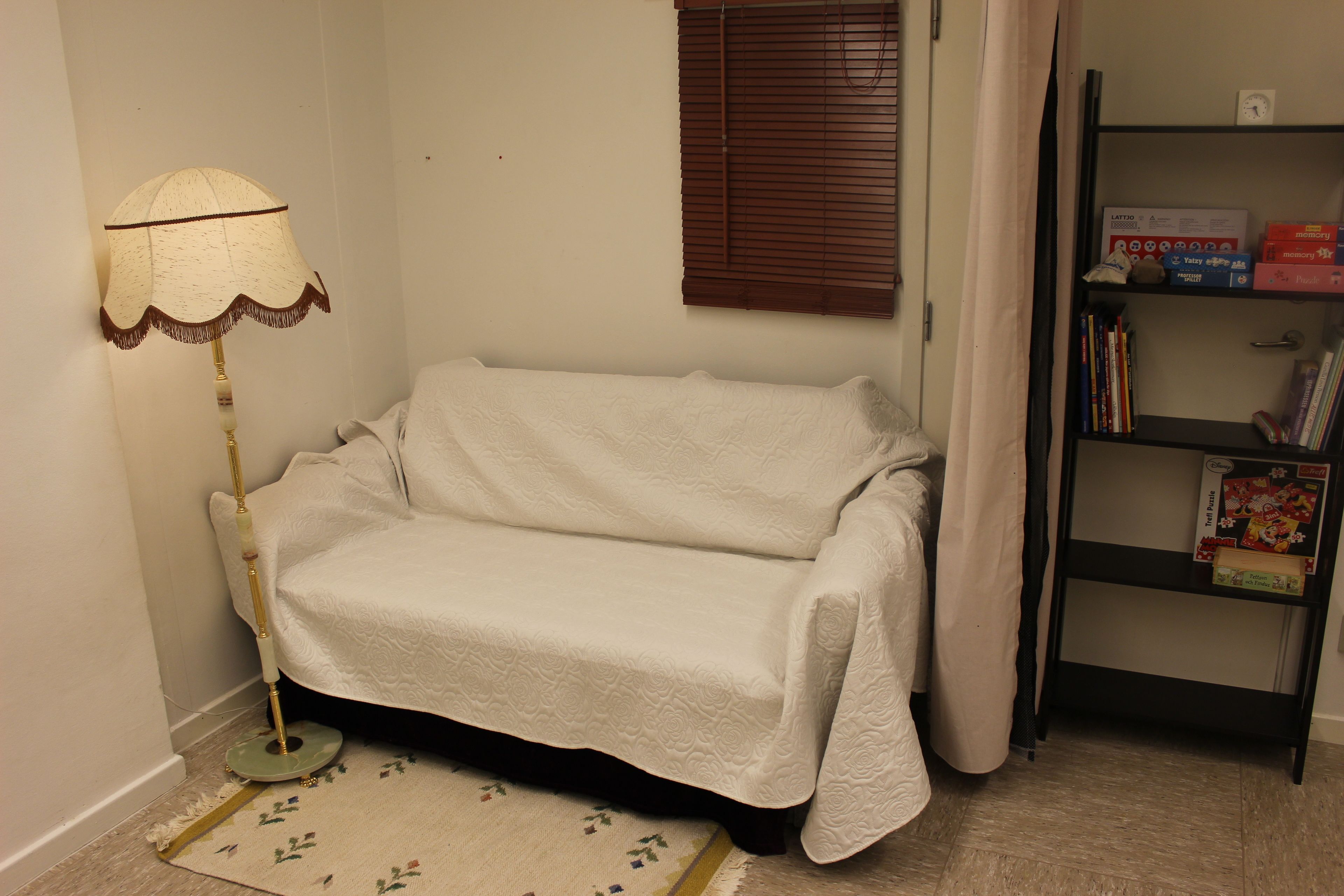 Una habitación con un sofá blanco, una lámpara de pie, una estantería y una ventana con persianas.