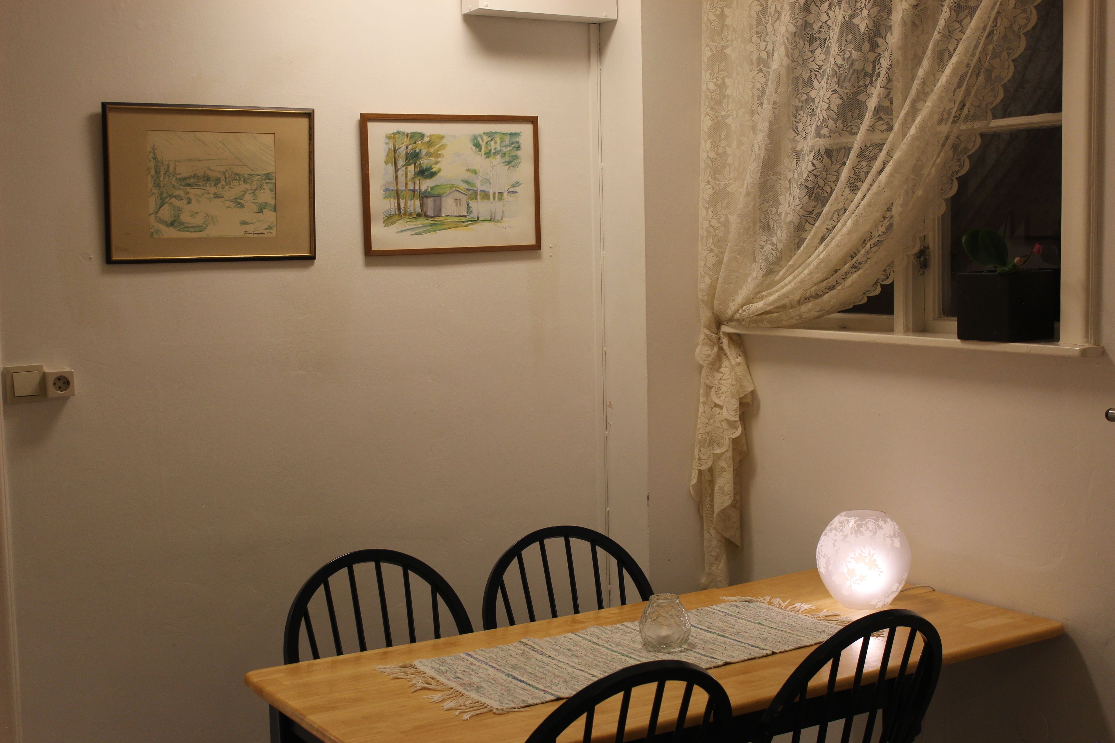 A dining area with a wooden table, four chairs, a vase, and two framed artworks on the wall.