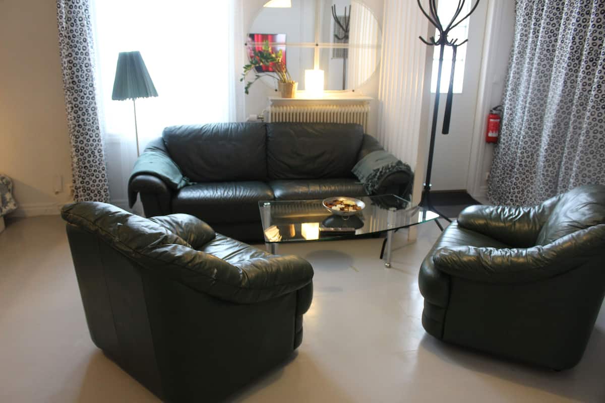 A living room with a black leather sofa, two armchairs, a glass coffee table, a lamp, a radiator, and a fire extinguisher.
