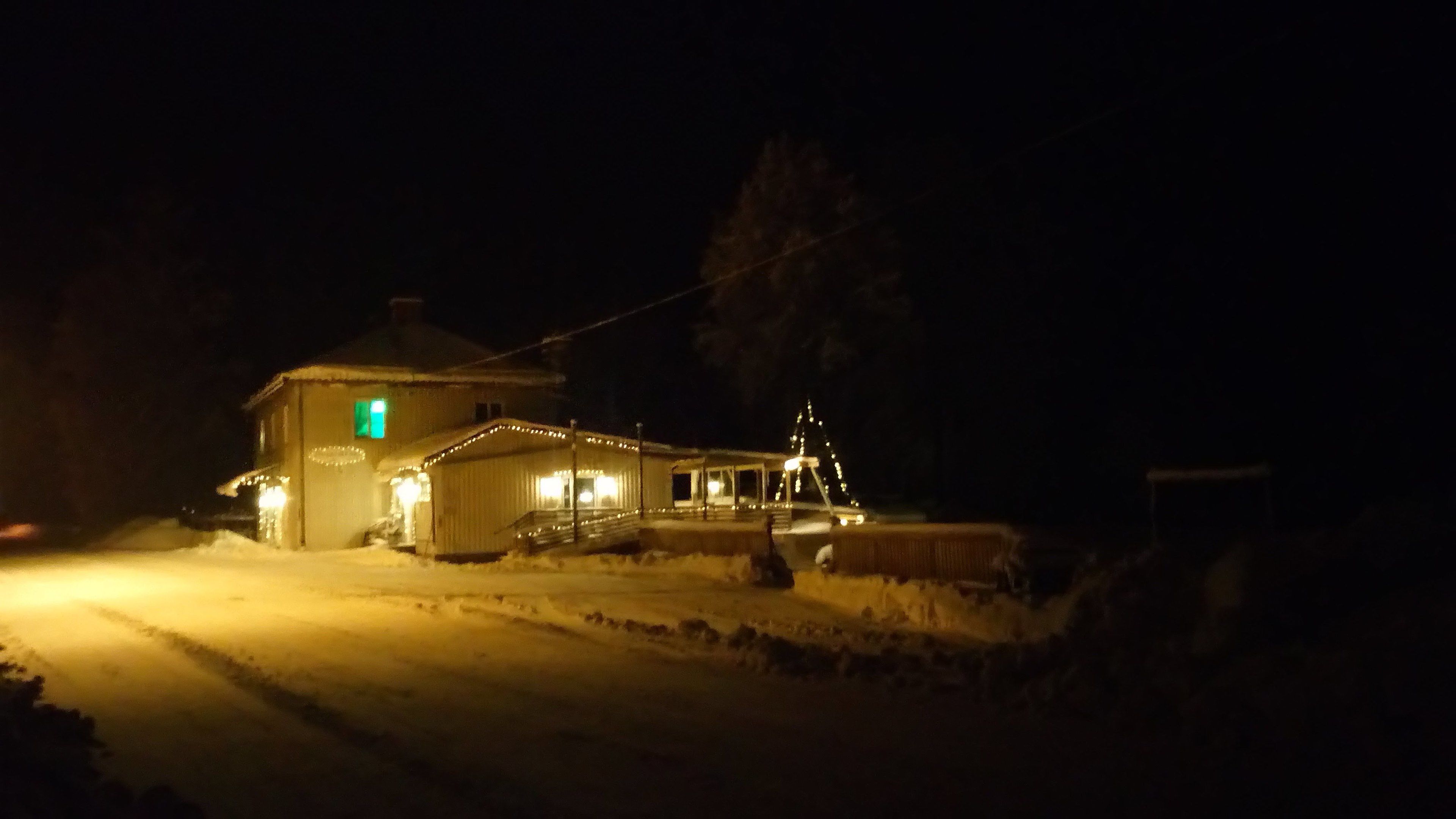 Un bâtiment recouvert de neige, avec une fenêtre illuminée de lumière verte, un porche abrité et un sol enneigé.