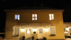 Une maison en bois à deux étages, dotée d’un porche, de fenêtres illuminées et d’une lune dans le ciel.