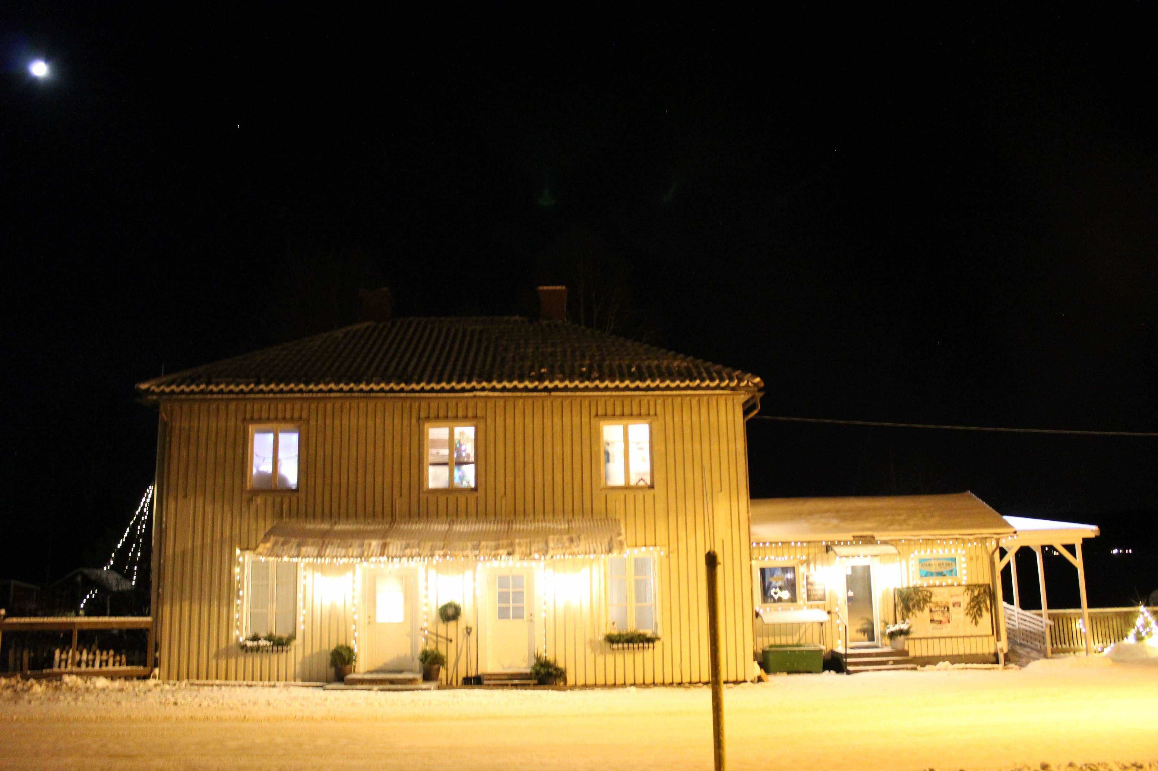 A two-story wooden building with a porch and a covered area, illuminated by interior lights.