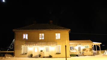 A two-story wooden building with a porch and a covered area, illuminated by interior lights.