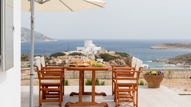 A wooden table with two chairs and a white umbrella, overlooking a coastal view with a white building and the sea. - Kimolos Houses (Kimolos)