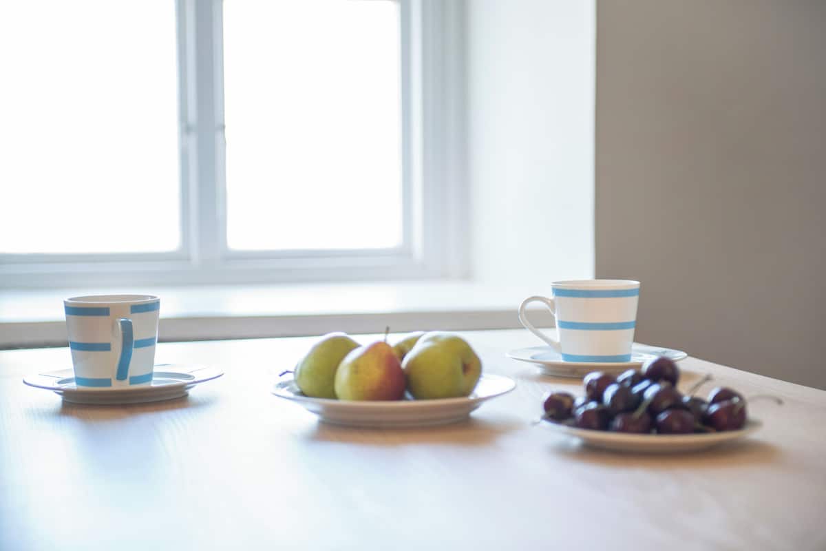 A table with a plate of green apples and a plate of cherries, two cups with blue stripes, and a window in the background.