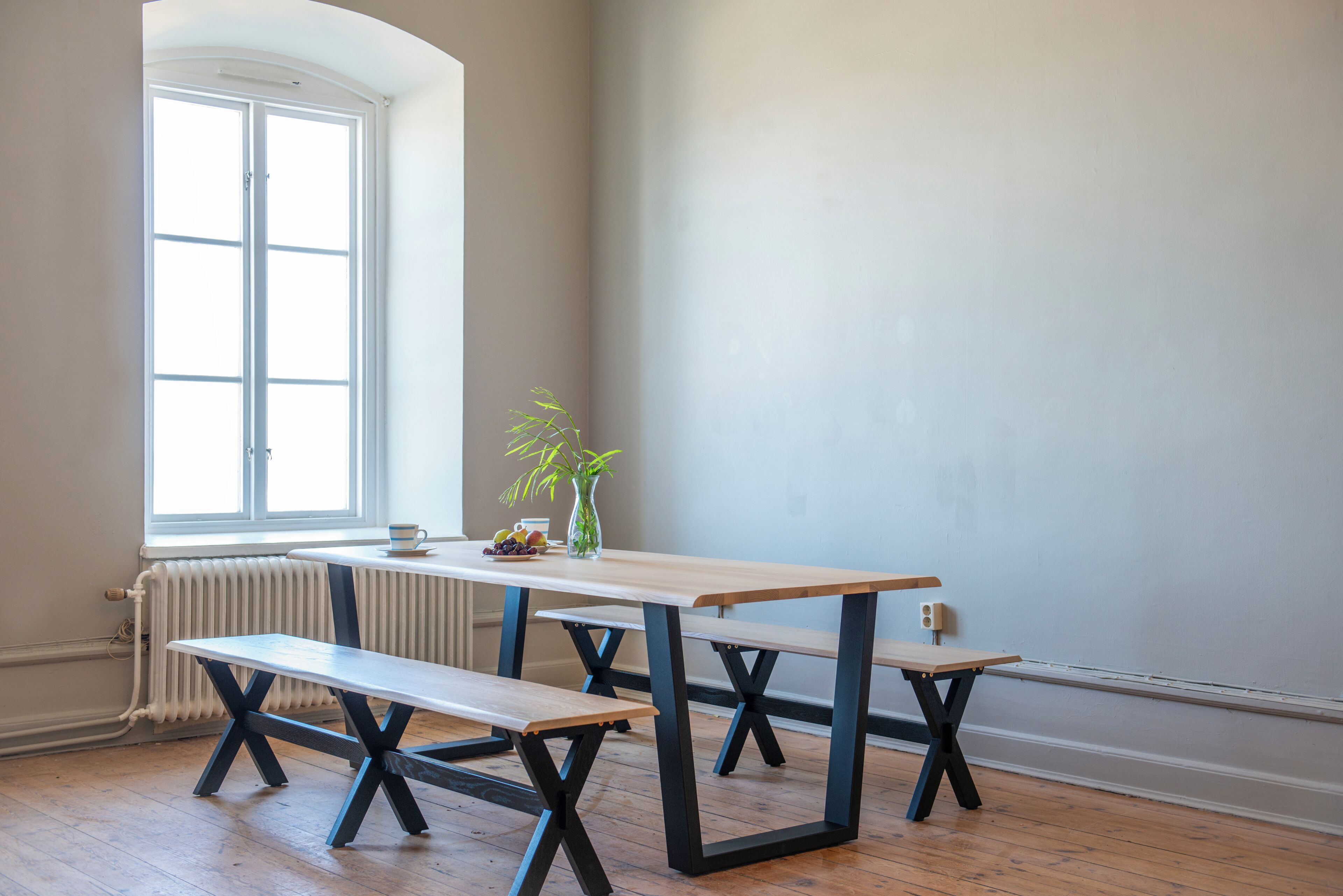A room with a wooden table and benches, a radiator, a window, and a vase with a plant.