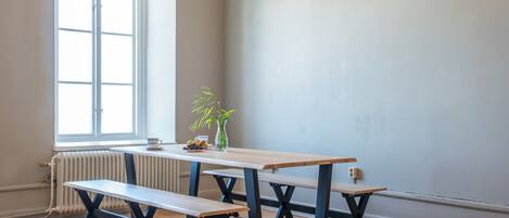 A room with a wooden table and benches, a radiator, a window, and a vase with a plant.