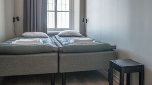 A twin bed with a grey headboard, a small bedside table, and a window with curtains. - STF Visby Hamn Vandrarhem (Visby)
