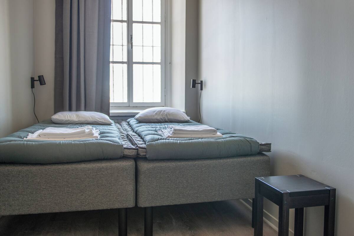 A twin bed with a grey headboard, a small bedside table, and a window with curtains.