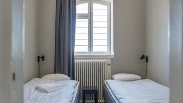 Habitación de hotel pequeña y con una cama, dos camas, una ventana con cortinas, un radiador y lámparas de noche.